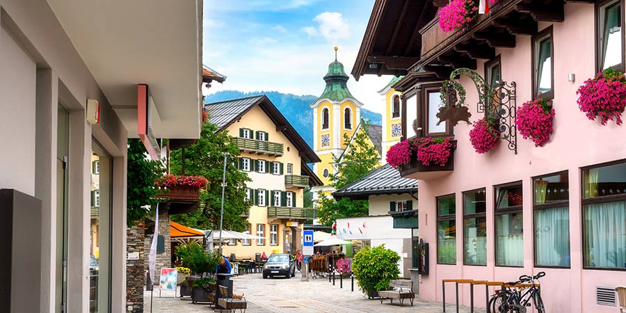 Scenic view of St. Johann in Tirol, a charming alpine town nestled in the mountains. Traditional colourful houses line the streets, with town a square at the centre and traditional church building. In the back are towering mountain peaks. 